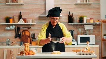 Woman baker using flour metallic sieve preparing homemade cakes. Happy elderly chef with bonete preparing raw ingredients to baking traditional bread sprinkling, sieving in the kitchen. photo