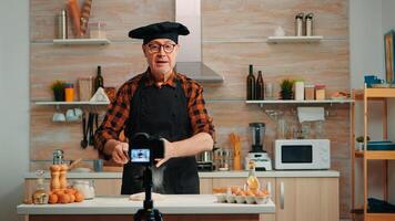 Cheerful elderly baker man filming cooking vlog in home kitchen. Retired blogger chef influencer using internet technology communicating, shooting blogging on social media with digital equipment photo