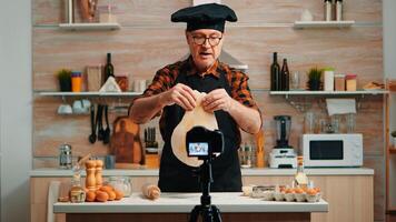 Baker using wooden rolling pin for dough in front video camera recording new cooking episode. Old blogger chef influencer using internet technology communicating on social media with digital equipment photo