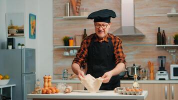 Old man with kitchen apron playing with bread dough at home smiling in front of camera. Retired elderly chef forming pizza countertop on a floured surface and kneading it with hands, in modern kitchen photo