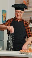 Experienced baker spreading flour in home kitchen for food preparation. Retired elderly chef with bonete and apron sprinkling, sieving sifting raw ingredients by hand baking homemade pizza, bread. photo