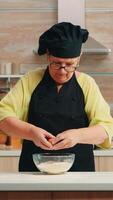 Woman baker cracking eggs into flour following traditional recipe in home kitchen. Retired elderly chef with bonete, mixing by hand, kneading in glass bowl pastry ingredients baking homemade cake photo