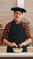 Old man wearing bonete while explaining food preparation recipe looking at camera. Retired blogger chef influencer using internet technology communicating on social media with digital equipment photo