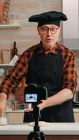 Cheerful elderly baker man filming cooking vlog in home kitchen. Retired blogger chef influencer using internet technology communicating, shooting blogging on social media with digital equipment photo