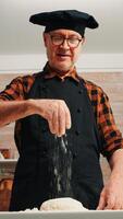 Senior man adding flour on dough by hand looking at camera smiling. Retired elderly chef with bonete and uniform sprinkling, sieving spreading rew ingredients with hand baking homemade pizza and bread photo