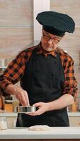 Baker with bonete and apron using metallic sieve preparing bakery products at home. Happy elderly chef with kitchen uniform mixing, sprinkling, sieving raw ingredients to baking traditional bread photo