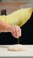 mujer con bonete y cocina delantal es ocupado con masa preparación. retirado mayor panadero con delantal, cocina uniforme aspersión, cernido, extensión harina con mano horneando hecho en casa Pizza y un pan. foto