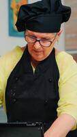 Cheerful retired chef in uniform spinning and tossing pizza dough throwing it up, forming on a floured surface and kneading it shaping. Skillful elderly baker wearing bonete cooking homemade cookies photo