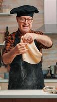 Old man with kitchen apron playing with bread dough at home smiling in front of camera. Retired elderly chef forming pizza countertop on a floured surface and kneading it with hands, in modern kitchen photo