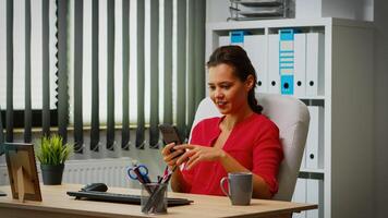 Hispanic lady chatting using smartphone in business office. Entrepreneur working in professional workspace, workplace in personal corporate company typing on mobile phone in front of computer photo