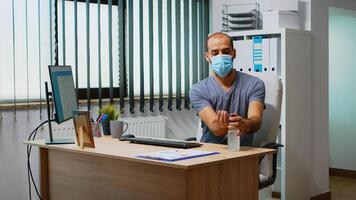 hombre vistiendo máscara y desinfectar manos en lugar de trabajo antes de mecanografía en teclado. empresario limpieza utilizando desinfectar alcohol gel en contra corona virus, trabajando en nuevo normal oficina lugar de trabajo en empresa foto