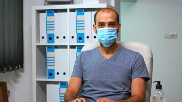 Man with mask working at computer and looking at camera sitting in office during pandemic. Entrepreneur in new normal personal workplace corporate writing on computer keyboard looking at desktop photo