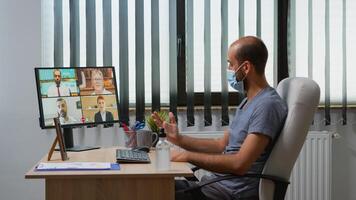 Freelancer making a video conference with face mask in modern office room. Entrepreneur working in new normal workplace having online meeting, webinar with remotely team using internet technology photo