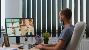 Business man with protective face mask talking on video call on pc while working in new normal office during coronavirus pandemic. Freelancer having online conference meeting using internet technology photo