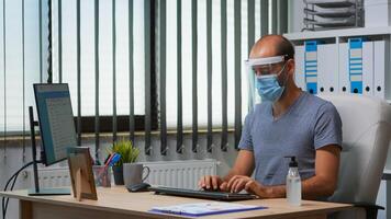 Man with protection mask and visor applying sanitizer gel rubbing hands before working at computer. Entrepreneur in new normal workplace disinfecting using antibacterial alcohol against corona virus. photo