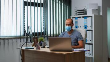 Man with mask having online meeting conference in modern office using laptop. Freelancer working in new normal office workplace chatting talking during virtual webinar, using internet technology photo