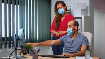 Coworkers with protection face masks working together in workplace during pandemic. Team in new normal office workspace in personal corporate company typing on computer keyboard looking at desktop photo