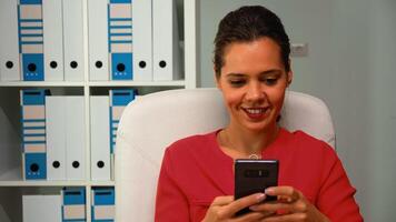 Woman browsing on smartphone smiling sitting on desk in modern office room. Hispanic entrepreneur working in professional workplace in corporate company typing on mobile phone in front of computer photo