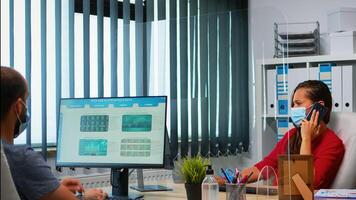 Woman with face mask talking on phone looking at desktop and analyzing the statistics. Freelancer working in workplace chatting with remotely team speaking on smartphone in front of computer photo