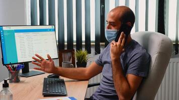 Manager wearing mask explaining on phone problem solution sitting on desk in office room. Freelancer working in new normal workplace speaking on smartphone with remotely colleagues in front of pc photo