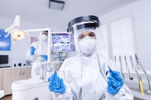 Patient pov of dentist in covid protectiv suit with her tools in dental office. Stomatolog wearing safety gear against coronavirus during heatlhcare check of patient. photo