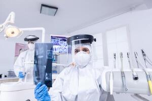 paciente pov de dental equipo explicando dental radiografía y diagnóstico porque dientes infección. estomatología especialista vistiendo protector materiales peligrosos traje en contra coroanvirus demostración radiografía. foto