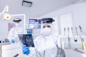 Patient pov of dentist examining patient teeth x-ray during covid gloabl pandemic. Stomatology specialist wearing protective hazmat suit against coroanvirus showing radiography. photo