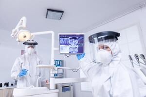 Patient pov of doctor pointing at stomatological monitor with dental x-ray. Stomatology specialist wearing protective suit against infection with coronavirus pointing at radiography. photo