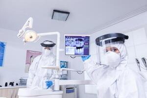 paciente pov de dentista con cara proteger explicando dental radiografía señalando a monitor. estomatología especialista vistiendo protector traje en contra infección con coronavirus señalando a radiografía. foto