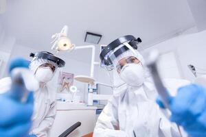 Dental team leaning over patient to work on patient gums dressed in hazmat suit. Stomatology team wearing safety equipment against coronavirus treating patient. photo