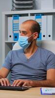 Businessman with medical protection face mask typing on computer in office room. Entrepreneur in new normal personal workplace corporate writing on pc keyboard looking at desktop photo