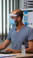 Man with protection mask and visor applying sanitizer gel rubbing hands before working at computer. Entrepreneur in new normal workplace disinfecting using antibacterial alcohol against corona virus. photo