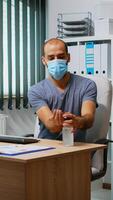 Man wearing mask and disinfecting hands in workplace before typing on keyboard. Entrepreneur cleaning using sanitize alcohol gel against corona virus, working in new normal office workplace in company photo