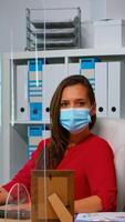 Woman with protection mask working and looking at camera sitting in front of pc. Employees in workspace in corporate company typing on computer keyboard looking at desktop respecting social distancing photo