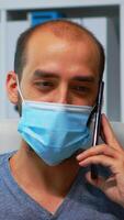 Portrait of man with protective mask talking on phone with partners in the office modern room during covid-19. Freelancer working in new normal workplace chatting writing speaking on smartphone. photo