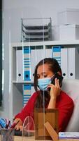 Woman with face mask talking on phone looking at desktop and analyzing the statistics. Freelancer working in workplace chatting with remotely team speaking on smartphone in front of computer photo