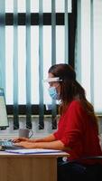 People with mask and visor working in new normal office checking reports and writing on computer. Coworkers in modern workplace respecting protection rules against covid virus using plexiglass. photo