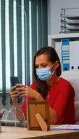 Woman with protective mask typing on phone while colleague working using clipboard respecting social distancing. Freelancer in new normal office chatting writing on mobile with internet technology photo