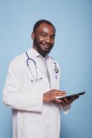 retrato de africano americano médico médico participación un tableta y mirando a cámara. sonriente masculino cuidado de la salud especialista avaro digital dispositivo mientras en pie en contra aislado azul antecedentes. foto