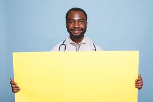 Smiling black doctor with stethoscope grasping large yellow banner for hospital advertising concept. Medical specialist presenting panel with empty copy space over isolated blue background. photo