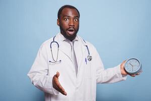 Medical practitioner looking surprised running out of time holding alarm clock while wearing lab coat and stethoscope. African american doctor panicking for being late for an appointment. photo