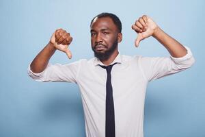 retrato de negro empresario en blanco camisa gesticulando dos pulgares abajo en desaprobación. fracasado masculino empresario demostración infeliz actitud gesto en pie en frente de aislado antecedentes. foto