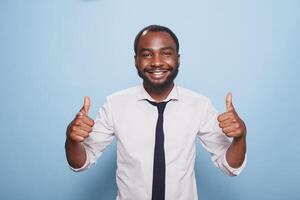 African American man is portrayed in front of the camera giving thumbs up with both his hands. Smiling black person spreading positivity, feeling joy and excitement and offering approval. photo