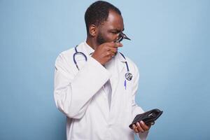 African american healthcare professional analyzing medical instrument used for ear examinations. Portrait of male doctor wearing lab coat and stethoscope while checking the otoscope in studio. photo