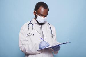 profesional médico vistiendo cara máscara y guantes es mirando a un portapapeles conteniendo paciente cartas y información. africano americano médico revisando médico investigación documentos. foto