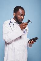 Male otologist of african american ethnicity is examining medical device used for human ear checkups. Shocked doctor inspecting an otoscope while dressed in a lab coat and stethoscope. photo