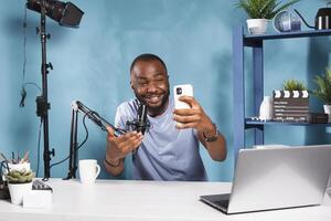 Cheerful blogger laughing while using smartphone for recording video. Smiling african american web content creator streaming on social media channel while holding mobile phone photo