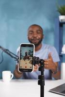 en línea flámula radiodifusión utilizando teléfono inteligente en trípode cerca arriba. africano americano joven hombre vlogger creando digital contenido y grabación vídeo para Blog en móvil teléfono foto