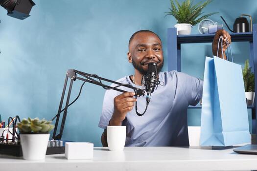 Vlogger announcing giveaway and showing shopping bag with gifts while broadcasting live. Smiling man blogger speaking to subscribers, promoting product and looking at camera photo