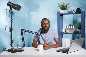 African american blogger recording vlog video and speaking in microphone portrait. Man streaming live on channel while creating digital content and looking at camera in home studio photo